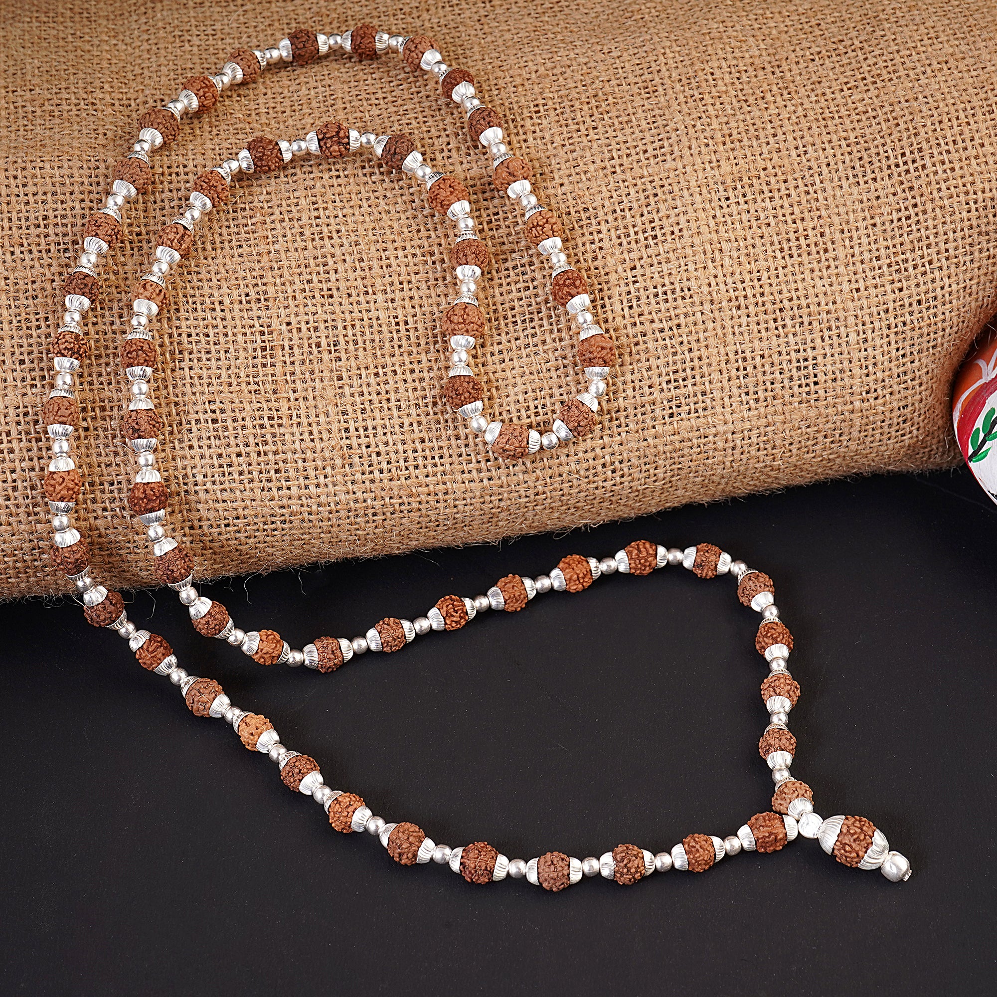 A beaded necklace featuring brown rudraksha beads and silver spacer beads, draped over a burlap roll on a black surface.