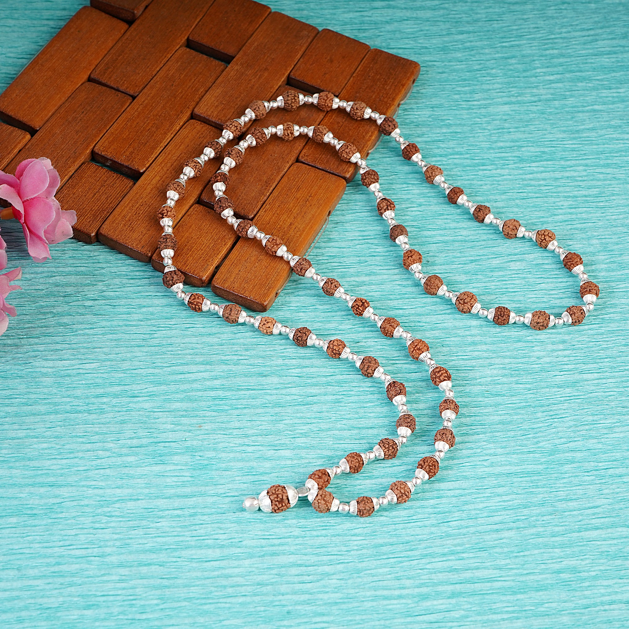 A beaded necklace made of brown textured seed beads and small white spacer beads, draped over a brown wooden tray on a turquoise textured background with a pink flower at the edge.