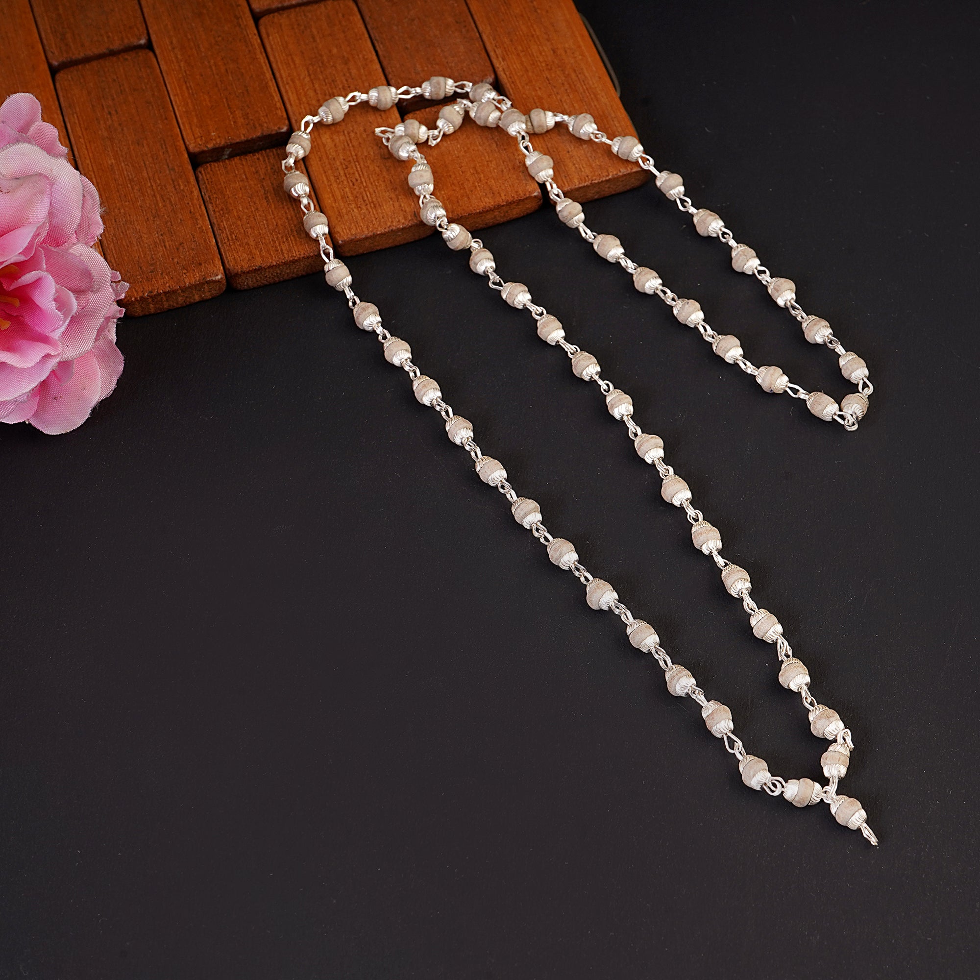 A silver-toned beaded necklace with irregular pearl-like beads, draped over a dark surface with a brown wooden block and a pink flower as props.