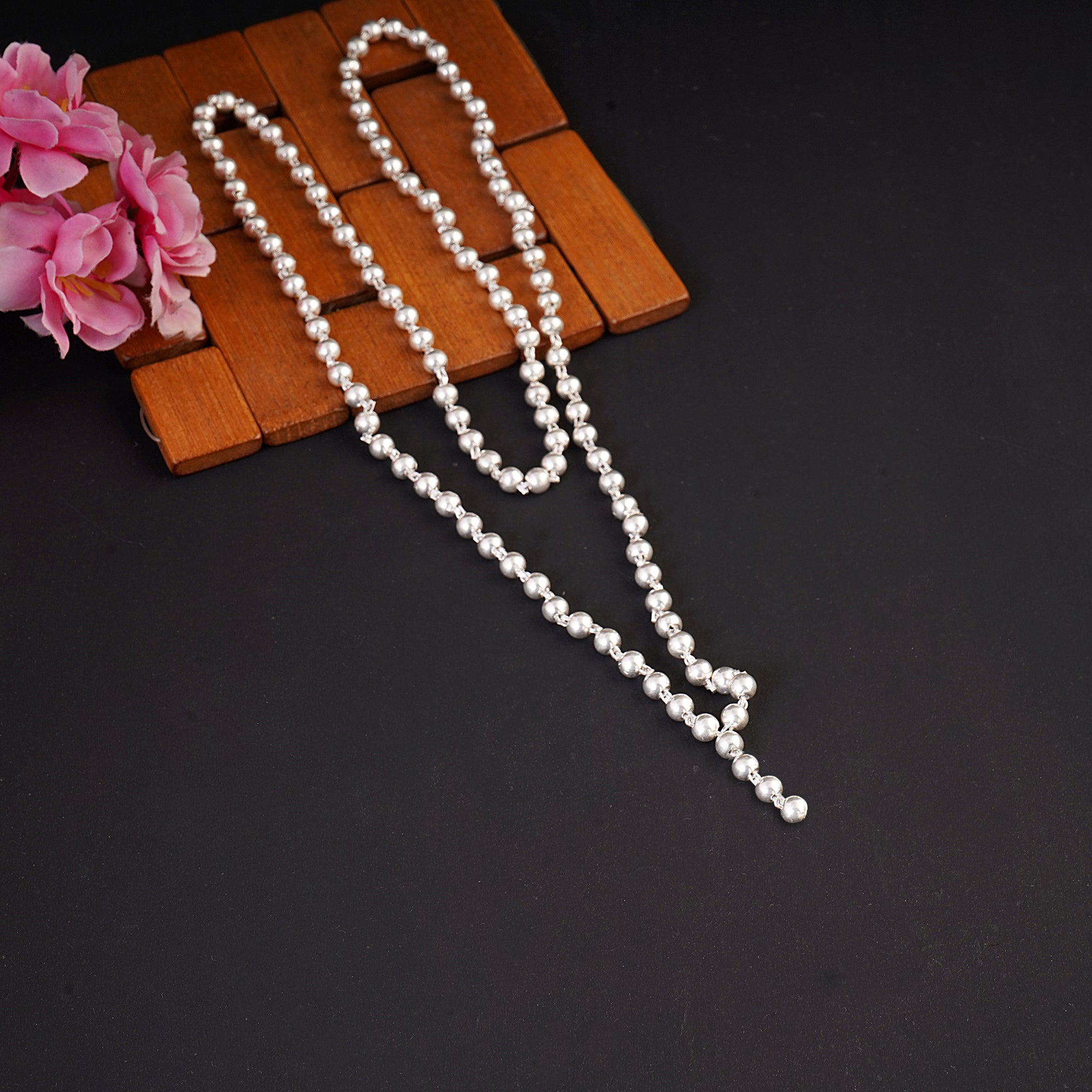A long strand of white faux pearls draped over a brown wooden block, with pink flowers in the top-left corner, set against a dark black background.